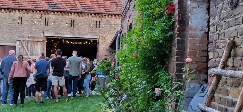 Menschenmenge vor einer Scheune mit blühenden Pflanzen im Vordergrund.