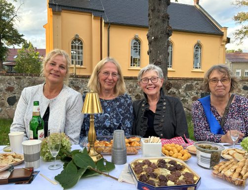 Vier Frauen lächeln an einem festlich gedeckten Tisch mit Speisen und Getränken.