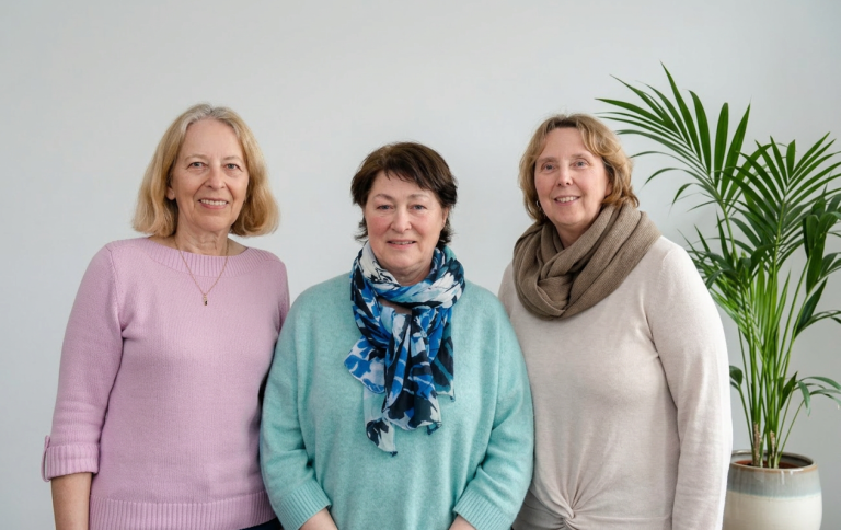 Drei Frauen lächeln, stehen nebeneinander vor einer hellen Wand mit einer Pflanze im Hintergrund.