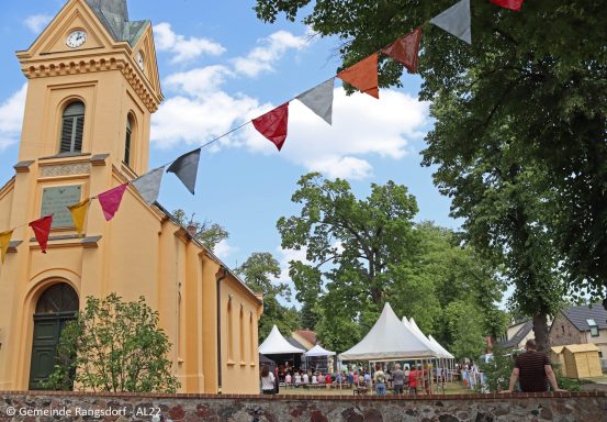 Bunte Wimpel über einem Festgelände mit einer Kirche im Hintergrund und Bäumen.