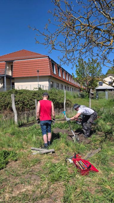 Zwei Personen arbeiten im Freien im Garten hinter einem Gebäude. Sonniger Tag.