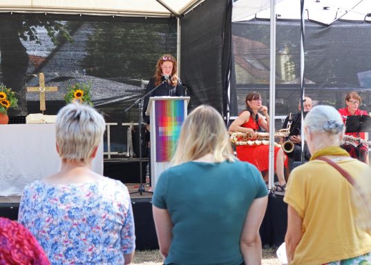 Eine Person spricht am Rednerpult vor einem Publikum, während Musikgruppen im Hintergrund spielen.