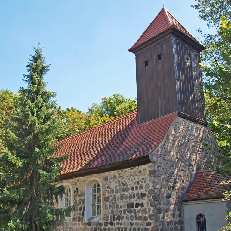 Kirche in Klein Kienitz umgeben von Bäumen und blauem Himmel.