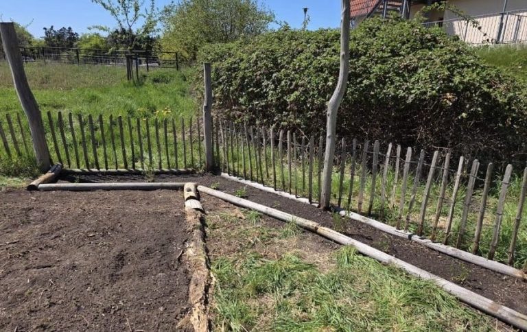 Zwei Personen stehen lächelnd neben einem bepflanzten Beet in einem sonnigen Garten.