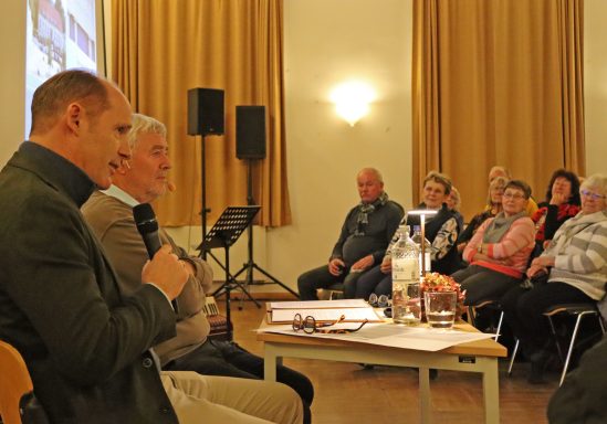 Zwei Männer halten einen Vortrag auf einer Bühne, Publikum sitzt aufmerksam und schaut zu.